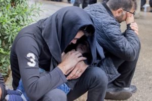 Two men crouched in the street, one holding the other as he covers his face