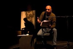 An actor seated at a table under a single light, his shadow thrown on the backdrop behind