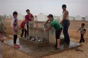 Children washing at an outdoor tap in front of rows of white tents