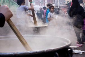 Hands stirring an enormous steaming cooking pot while a crowd waits behind