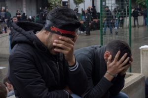 Two young men crouched with their hands to their faces, police standing behind