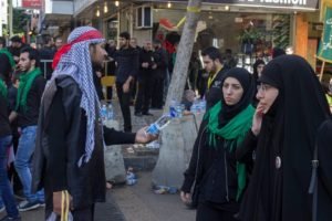 A man in a red keffiyeh handing out water to women during a street procession